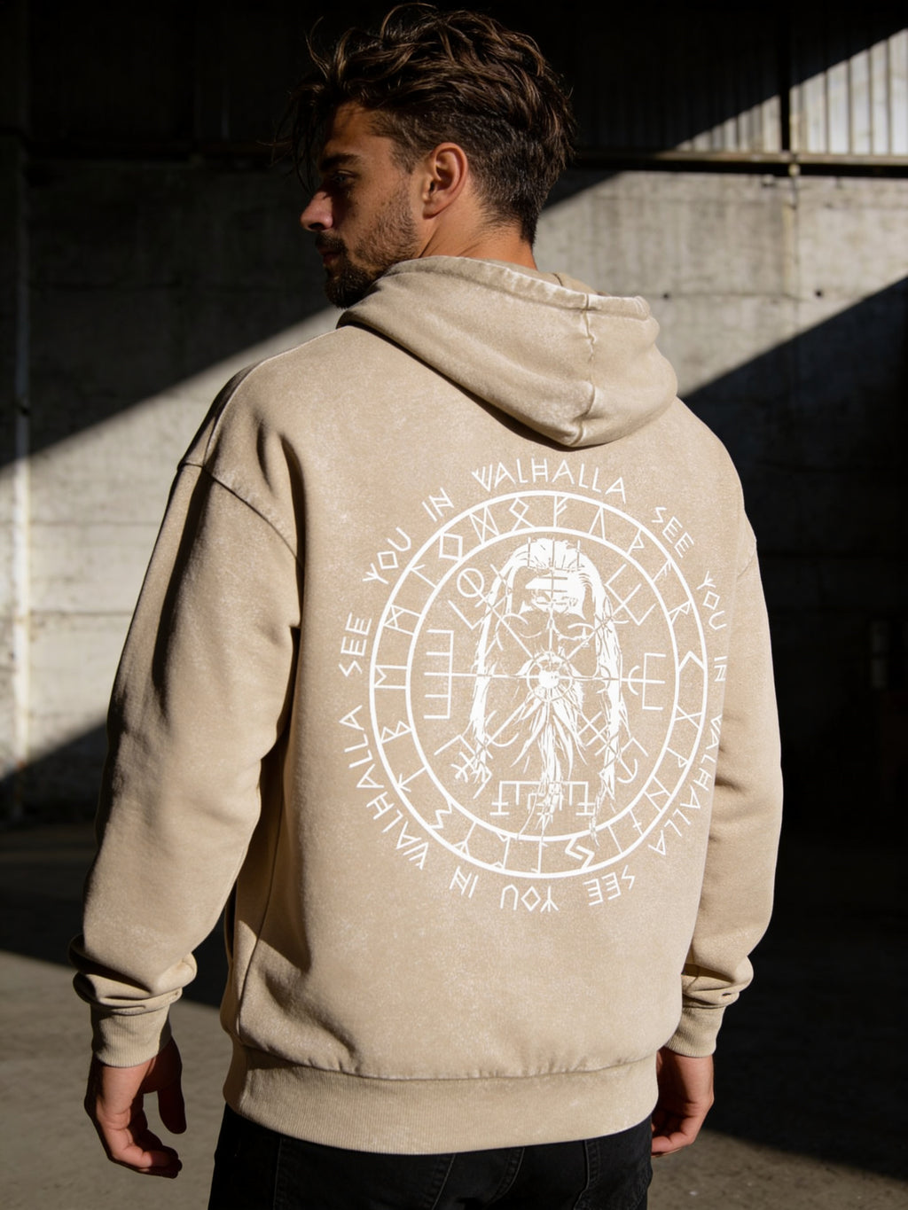 A man in a brown washed Oversize Hoodie Valhalla stands with his back to the camera in sunlight, showing a large circular Vegvísir design and “See you in Valhalla” printed on the back.