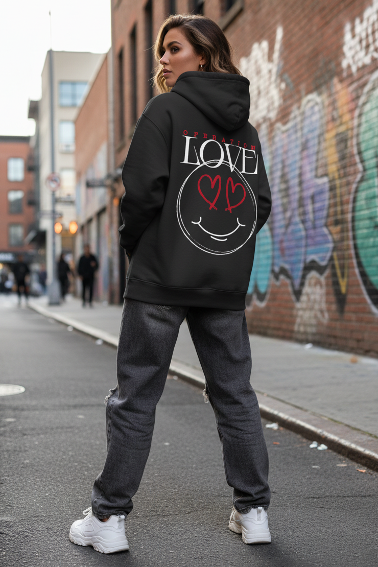Eine Frau steht auf einer von Graffiti gesäumten Straße und trägt den State Your Mind Oversize Hoodie Operation Love Black mit einem großen weißen Smiley-Gesicht mit roten Herzaugen und einem fetten LOVE"-Rückenprint, dazu graue Jeans und weiße Turnschuhe.