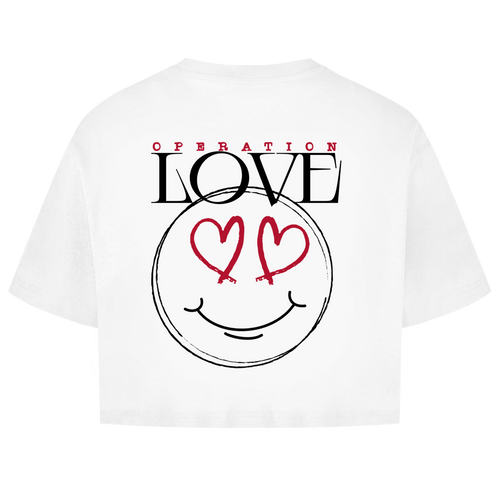 Das Oversize Crop Top Operation Love White von State Your Mind ist ein freches Streetwear-Stück für Damen, das mit dem Schriftzug "OPERATION LOVE" und einem großen schwarzen Smiley-Gesicht mit herzförmigen roten Augen als Rückenprint auf der Rückseite aufwartet.