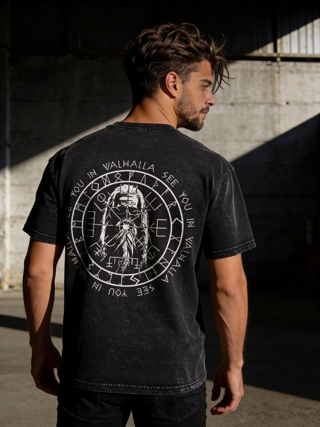 Ein Mann mit dunklem Haar und Bart steht im Sonnenlicht. Er trägt ein Printegy Oversize Shirt Valhalla Black washed mit einem kreisförmigen SEE YOU IN VALHALLA-Motiv mit nordischen Motiven und einer Wikingerkompass-Krieger-Grafik auf dem Rücken.