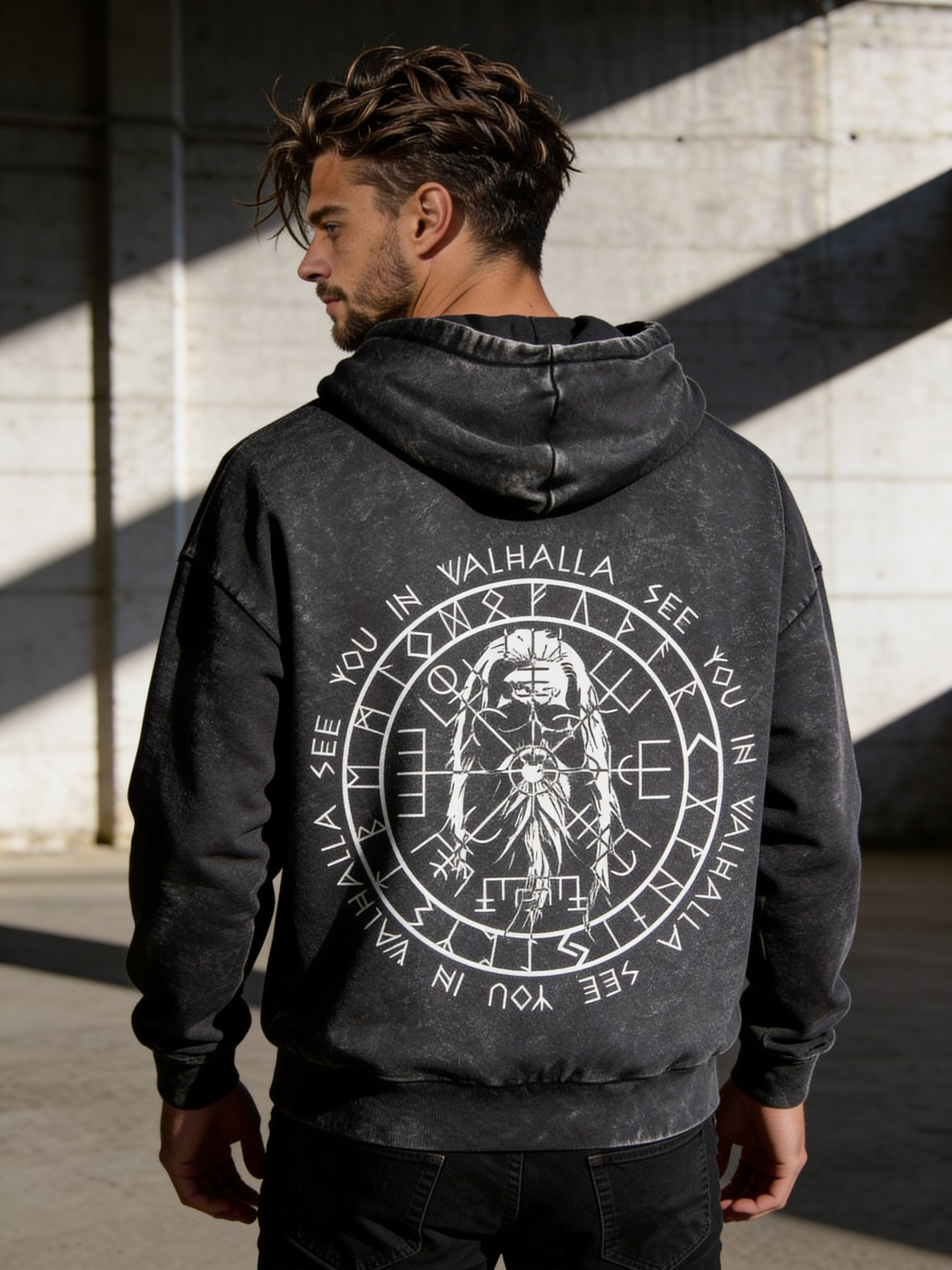 Ein Mann mit gewelltem braunem Haar trägt den State Your Mind Oversize Hoodie Valhalla Black washed mit nordischen Runen, einem Walkürenmotiv, einem Vegvísir-Symbol und dem Schriftzug See you in Valhalla" und steht in einer sonnenbeschienenen Industriehalle.